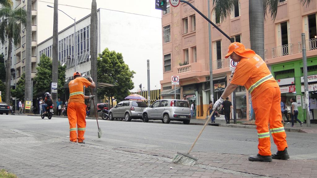 Duas mulheres garis varrendo a calçada de uma grande avenida da cidade.