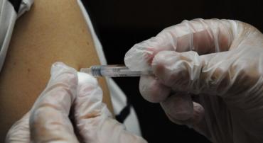 Mãos enluvadas aplicam vacina em braço de cidadão. 