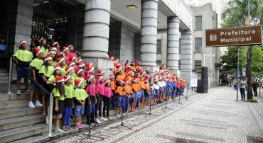 Cerca de 400 estudantes de escolas municipais vestindo gorrinhos de Natal, cantam na entrada da Prefeitura. 