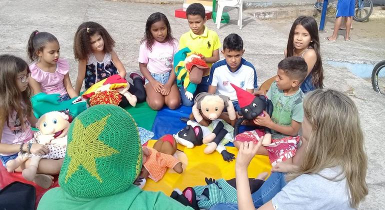Nove crianças manuseiam város bonecos na praça. Atividade faz parte do Programa Escola Aberta.