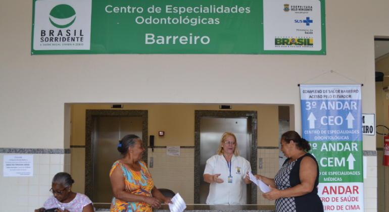 Atendente do Centro de Especialidades Odontológicas Barreiro Atende a duas mulheres de pé, à esquerda, uma outra mulher está sentada. 