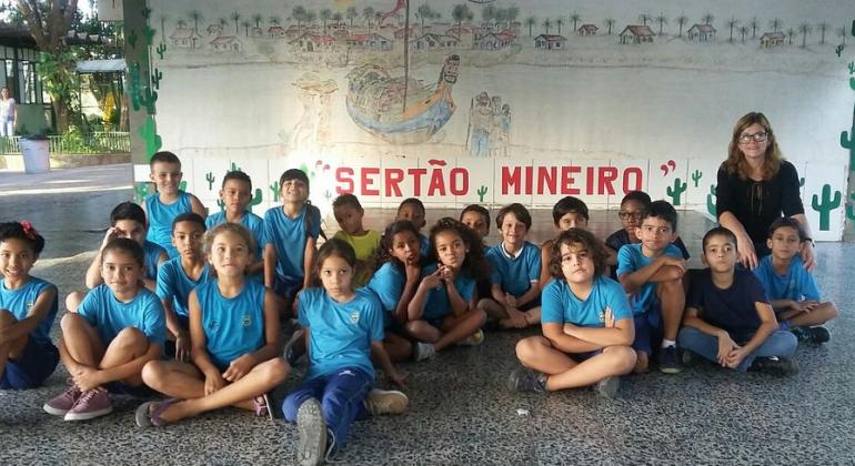 Dezesseis alunos sentados no chão da Escola Municipal Minervina Augusta, acompanhados de professora. Ao fundo um cartaz com os dizeres: "Sertão Mineiro".