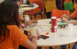 Três crianças sentada em volta de uma mesa branca redonda, vestindo camisetas alaranjadas fazem trabalhos manuais usando materiais recicláveis como papelão, copo de plástico e outros 