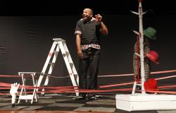 Comediante segura celular vermelho em palco com adereços como escada, cabideiro e banco brancos. Foto: Divulgação