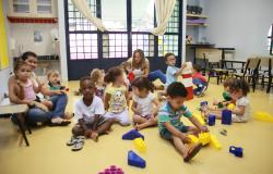 Em sala de aula, cerca de 10 crianças sentadas empilham blocos ao lado de duas professoras