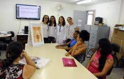 Mulheres do centro de saúde mostram desenho do corpo feminino, com destaque para os órgãos reprodutivos, para cidadãs sentadas: quatro mulheres estão de pé, e quatro estão sentadas. 