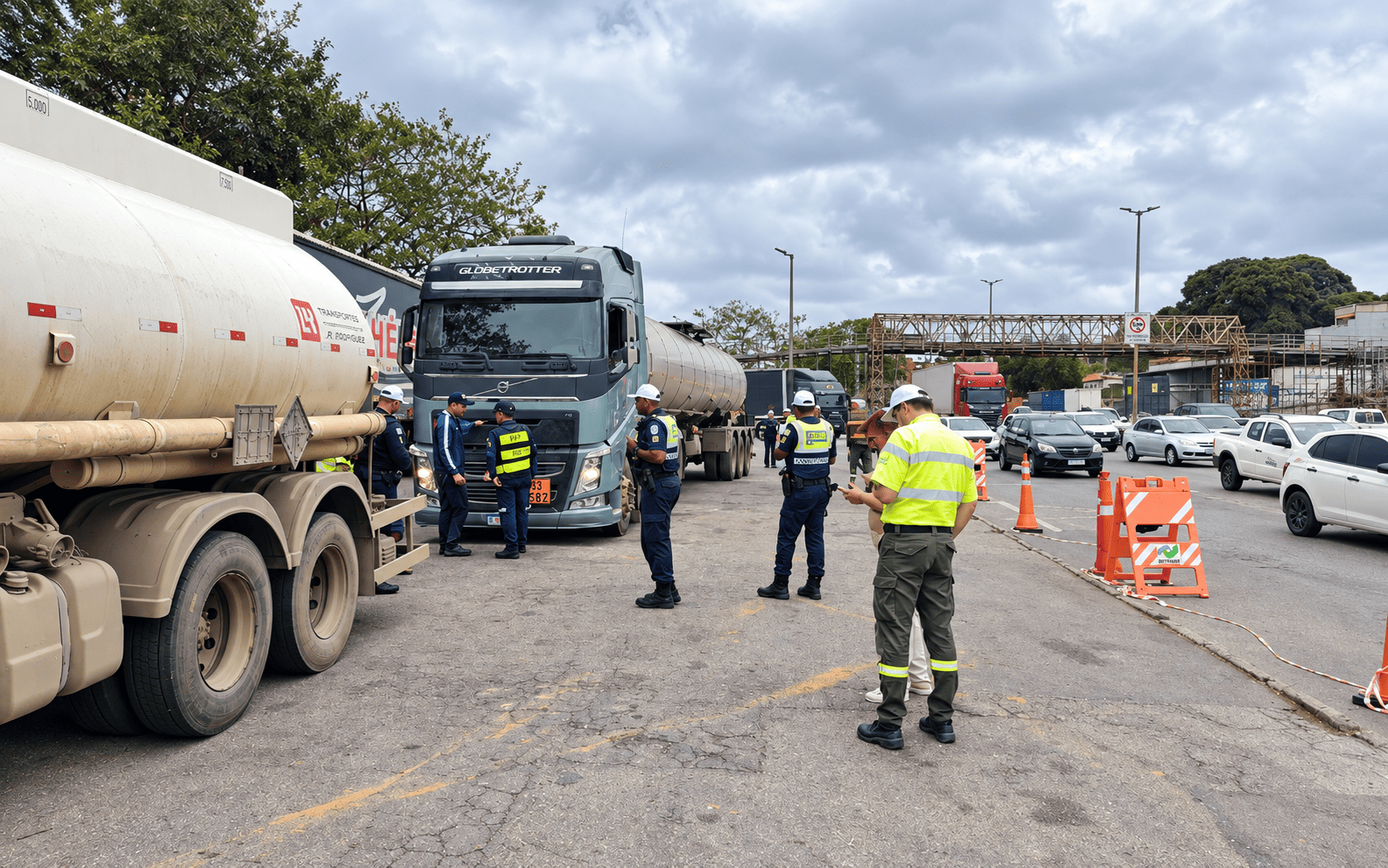 PBH faz nova operação conjunta com a ANTT no Novo Anel para fiscalizar veículos que transportam cargas perigosas A imagem mostra uma operação de fiscalização rodoviária a camiões cisterna. Diversos agentes fardados e com coletes refletores inspecionam os veículos e documentos no local, sinalizado com cones e barreiras cor-de-laranja.