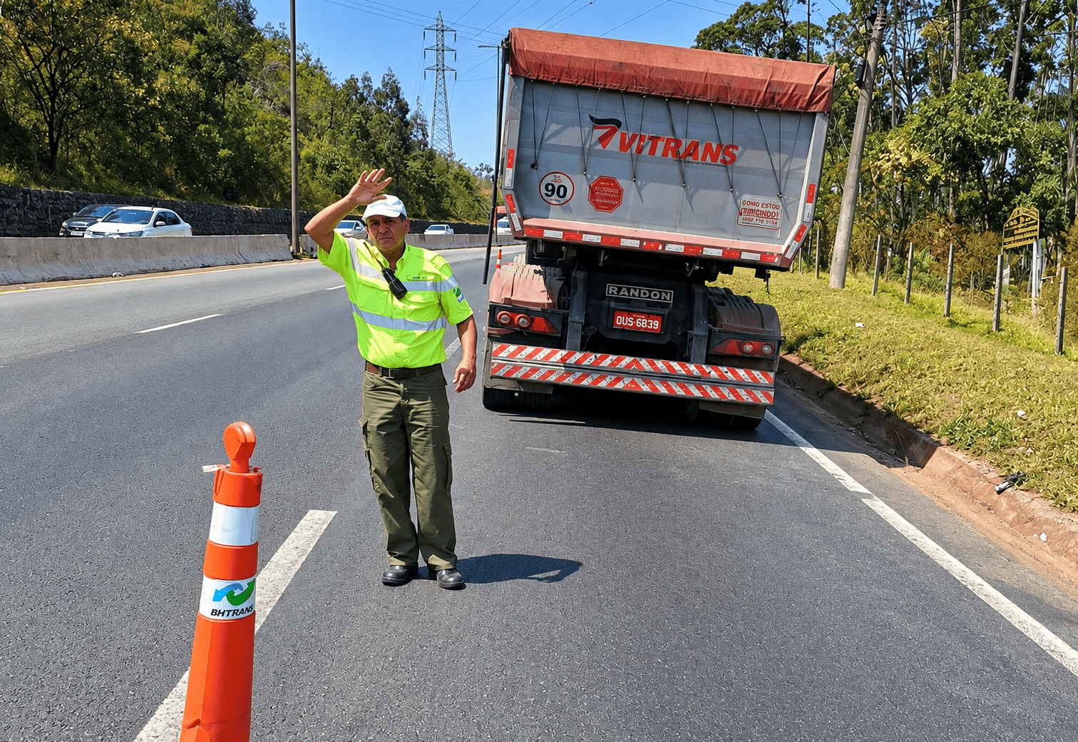 PBH faz nova operação conjunta com a ANTT no Novo Anel para fiscalizar veículos que transportam cargas perigosas A imagem mostra um agente de trânsito em pé numa estrada, a acenar com a mão para orientar o tráfego junto a um cone de sinalização. Ao seu lado, está parado na berma um camião de mercadorias com a traseira visível.