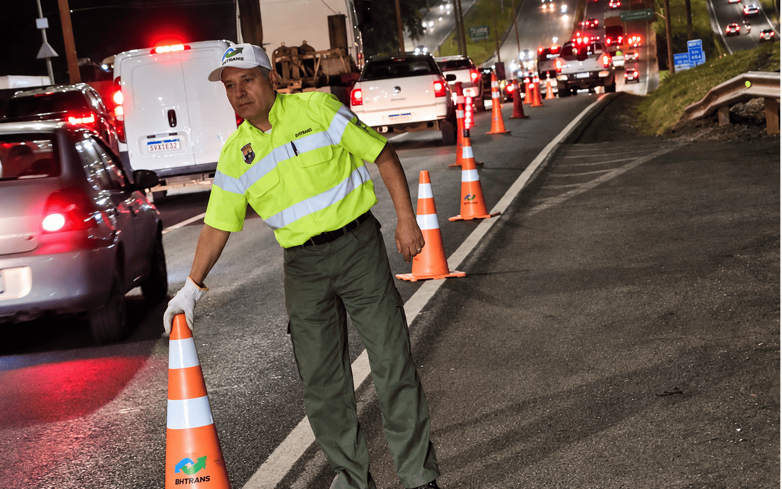 PBH faz nova operação conjunta com a ANTT no Novo Anel para fiscalizar veículos que transportam cargas perigosas A imagem mostra um agente da BHTRANS com colete refletor a posicionar cones de sinalização Laranja para organizar o trânsito ou montar um ponto de fiscalização noturno, numa via movimentada com vários veículos ao fundo.