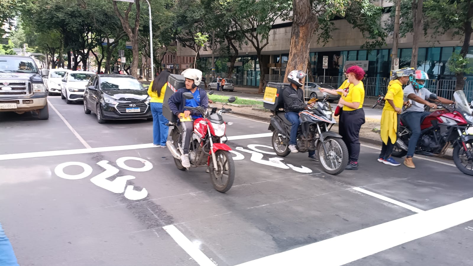 Dia Nacional do Motociclista será celebrado na próxima segunda-feira (27) com ação educativa na Afonso Pena Motociclista parados na faixa exclusiva. Mulheres com blusas amarelas entregando panfletos para esses motociclistas. Ambiente aberto, no meio da rua, em local com semáforo no vermelho.