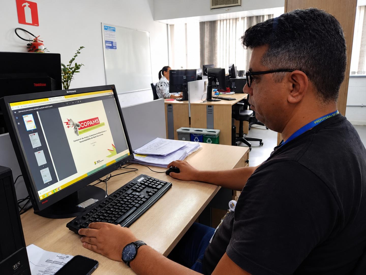 PBH convoca 47 assistentes de apoio administrativo aprovados em processo seletivo Homem sentado à mesa de um escritório, usando óculos, camisa preta e crachá da Prefeitura. Ele trabalha ao computador, com uma apresentação aberta na tela e documentos sobre a mesa. Ao fundo, há outras estações de trabalho, computadores e uma colega em atividade.