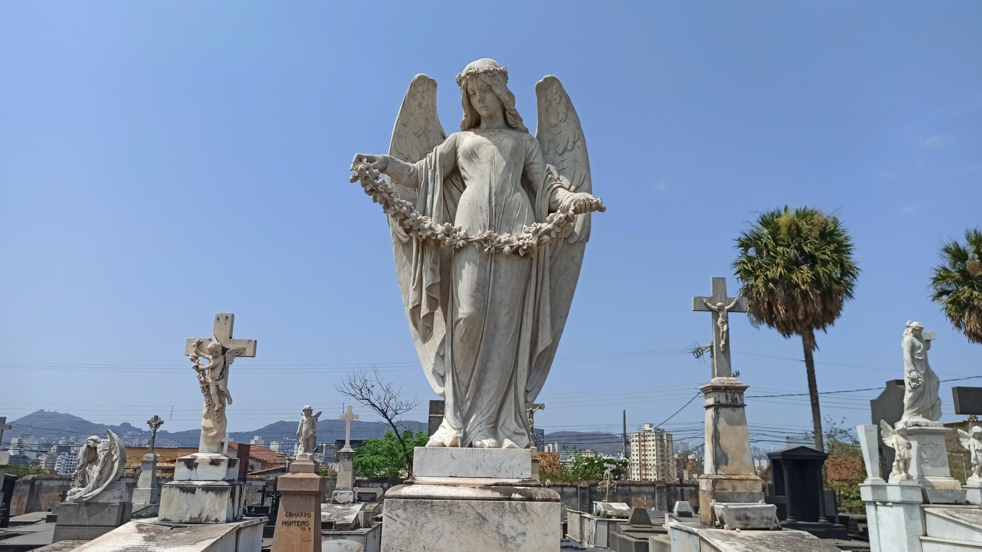 Prefeitura de BH oferece serviço de sepultamento gratuito para famílias em situação de vulnerabilidade A imagem mostra esculturas e túmulos do Cemitério do Bonfim, em Belo Horizonte, sob um céu azul e ensolarado. No centro, destaca-se uma grande escultura de mármore branco representando um anjo de asas abertas, cabelos longos e vestes esvoaçantes. Com os braços estendidos, a figura segura uma guirlanda de flores. Ao redor, há túmulos, crucifixos e outras esculturas religiosas, incluindo anjos e figuras humanas. Ao fundo, aparecem palmeiras, prédios e montanhas que compõem a paisagem urbana de Belo Horizonte.