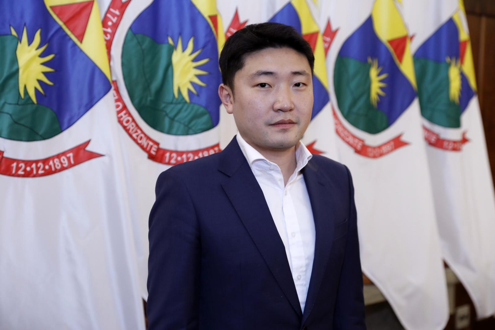 Cheng Qu, Secretário de Relações Institucionais, de terno, olhando em direção a câmera, e ao fundo, bandeiras de Belo Horizonte 