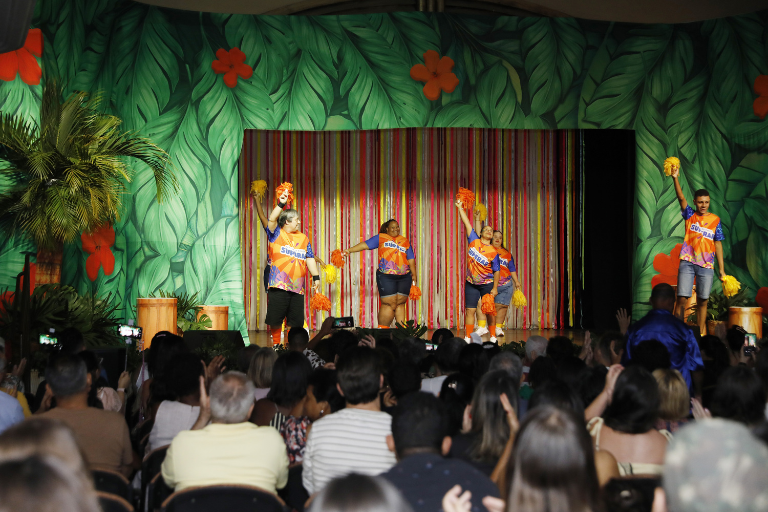 Registro da 18ª edição do Festival de Dança do Programa Superar, realizada em 2025. No palco, cinco participantes vestem camisetas coloridas em tons de laranja, azul e rosa, com a identificação do programa. Durante a apresentação, dançam segurando pompons amarelos e alaranjados. O cenário é decorado com grandes folhas verdes, flores e fitas coloridas. À frente do palco, o público acompanha a apresentação, aplaude e registra o momento com celulares.