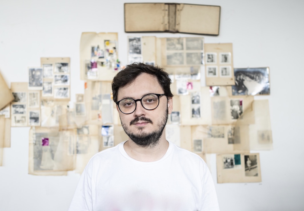 Museu de Arte da Pampulha recebe instalação audiovisual que passeia pelo universo da música caipira Imagem do artista Benedito Ferreira, um homem de óculos de armação preta e camiseta branca, de barba e bigode, posando diante de uma parede coberta por fotografias, documentos e imagens antigas organizadas em painéis.