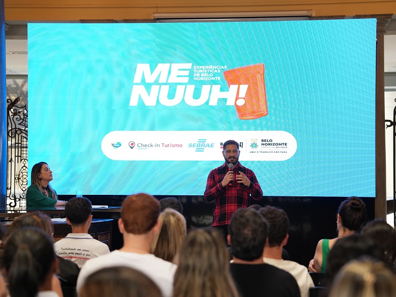 O presidente da Belotur, Eduardo Cruvinel, fala durante o lançamento da segunda etapa do "Menuuh! – Experiências Turísticas de Belo Horizonte". Ao fundo, o painel apresenta a identidade visual do projeto,