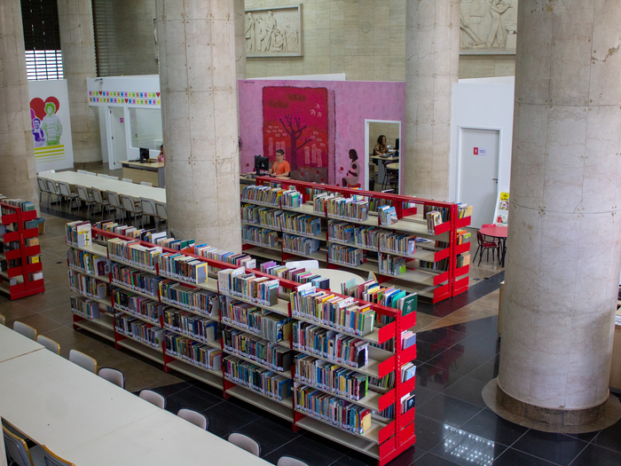 “Férias na Biblioteca” reúne literatura, teatro e oficinas para todas as idades Estantes na horizontal, na cor vermelha, com vários livros coloridos. As estantes estão entre pilastras na cor marfim