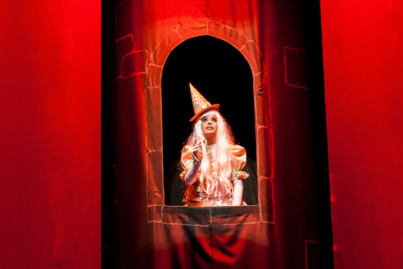 Espetáculo infantil no Teatro Marília fala sobre o respeito às diferenças No centro, uma atriz interpretando olha para cima com expressão esperançosa, emoldurada pela janela oval de uma torre escura. Ela usa chapéu pontudo bege decorado, peruca longa e branca, e vestes em tons claros e dourados. A torre está centralizada contra um fundo e iluminação com fortes tons avermelhados nas laterais.