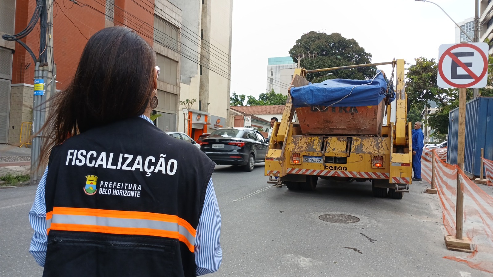 Fiscalização combate instalação irregular de caçambas com mais de 1,7 mil vistorias neste ano em BH Agente de fiscalização da Prefeitura de Belo Horizonte, de costas, usando colete de trabalho na cor azul, com listra alaranjada, acompanha caminhão com caçamba em uma rua com área de obras isolada.