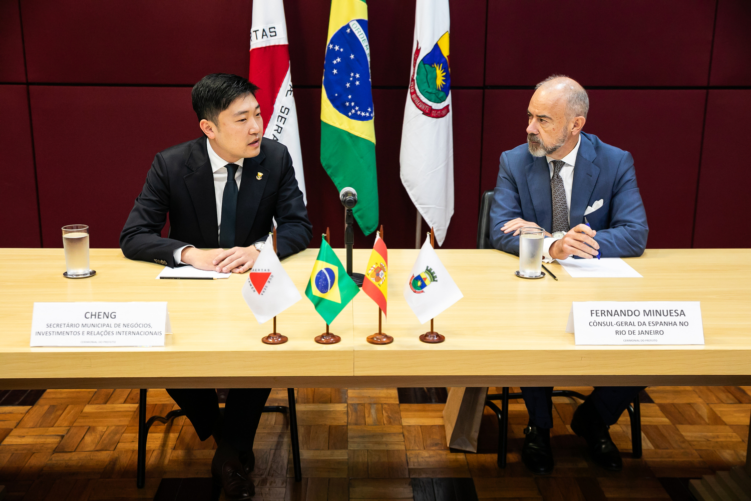 Belo Horizonte e Espanha debatem parcerias para atração de investimentos e cooperação internacional Foto de Qu Cheng é o secretário municipal de Negócios, Investimentos e Relações Internacionais, e o cônsul-geral da Espanha, Fernando Fernández-Arias Minuesa, sentados à mesa, com as bandeiras de Minas, Brasil, Espanha e PBH