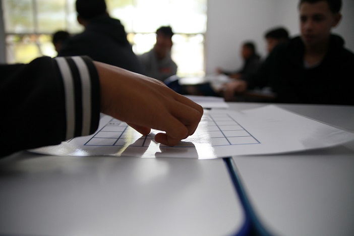 Escolas municipais participam das Jornadas TOP com desafios, jogos e experiências práticas Imagem mostra a mão de um aluno apontando para um papel que está sobre uma bancada clara e lisa. Ao fundo há imagens distorcidas de pessoas e de uma janela pela qual entra um pouco de luz natural.