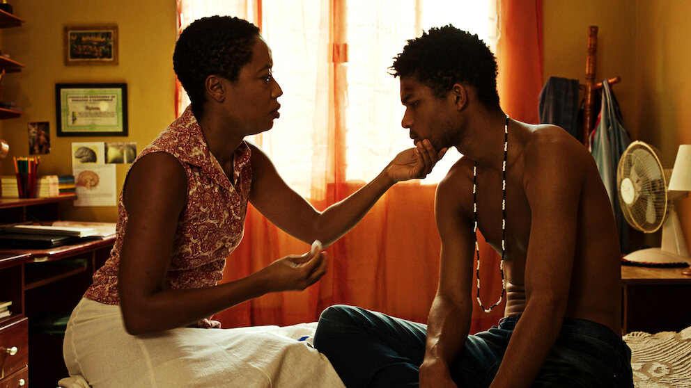 Cine Santa Tereza exibe filmes brasileiros na mostra “Especial Cinema Negro” Cena do filme "M8 - Quando a morte socorre a vida".Uma mulher negra de cabelo curto, vestindo uma regata estampada e saia branca, ergue suavemente o queixo de um jovem negro, enquanto segura um pedaço de algodão na outra mão. O rapaz está sem camisa, usa um colar de contas e fica sentado na cama olhando para ela com uma expressão pensativa. Eles estão em um quarto iluminado por uma luz quente e alaranjada.
