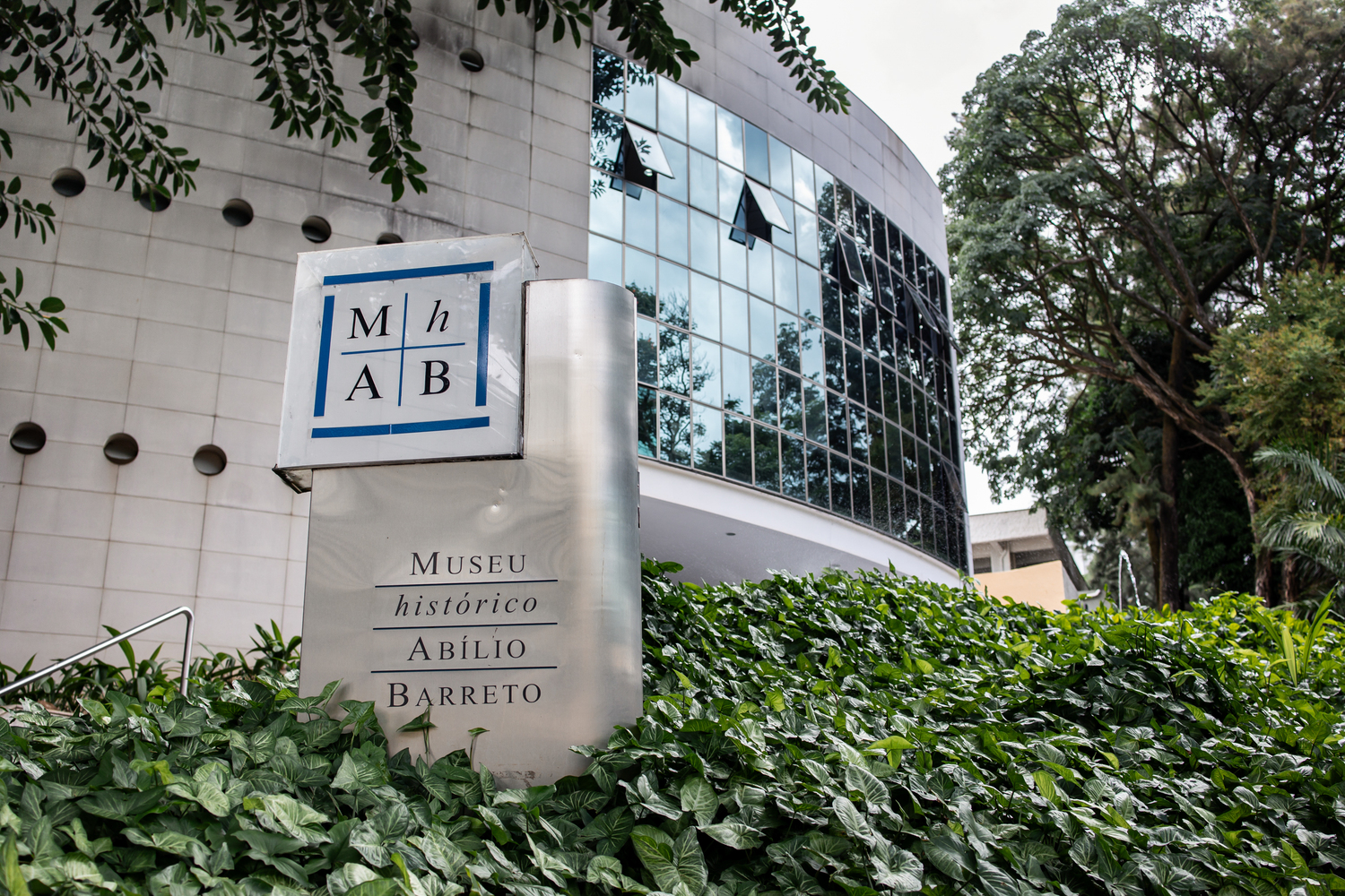 Prefeitura de BH seleciona Organização da Sociedade Civil para o Projeto “Museus BH” Fotografia em plano médio da fachada do Museu Histórico Abílio Barreto. Em primeiro plano, no canto inferior esquerdo, destaca-se um tótem em metal com uma placa decorativa onde se lê a sigla "MHAB" dividida em quatro quadrantes azuis e a inscrição "Museu Histórico Abílio Barreto". O totens surge do meio de uma folhagem verde e densa.
