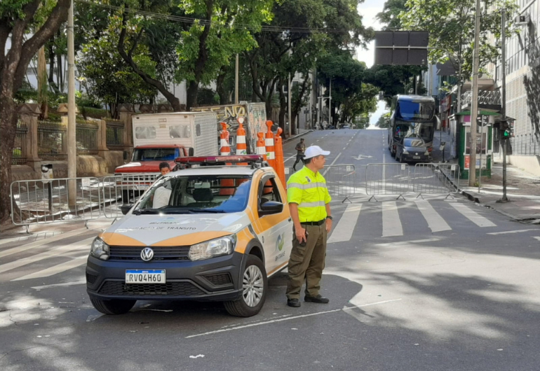 BHTrans realiza operações de trânsito para eventos na capital neste final de semana com Linha 55 Agente de trânsito usando uniforme da BHTrans, nas cores verde e amarelo, com boné branco, ao lado de veículo usado em operação de trânsito, parado em via de BH