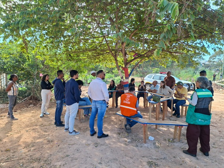 PBH e moradores promovem mutirão de limpeza e conscientização ambiental no Novo Aarão Reis Serviores da prefeitura, usando uniforme laranja e verde, conversando com pessoas sentadas em bancos colocados ao lado de árvores e viatura da guarda municipal