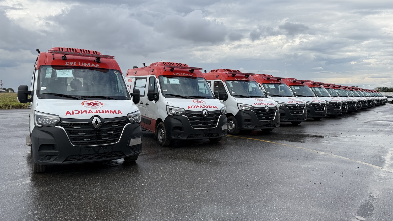 Belo Horizonte recebe 17 novas ambulâncias para renovação da frota do Samu A fotografia mostra uma fileira de novas ambulâncias brancas e vermelhas do SAMU 192, alinhadas num pátio de asfalto molhado sob céu nublado. A van em primeiro plano exibe em destaque o logotipo "SAMU 192", o símbolo da Estrela da Vida, a inscrição "Serviço de Atendimento Móvel de Urgência", a bandeira do Brasil e a marca do SUS, enquanto os outros veículos do mesmo modelo se estendem em perspectiva ao fundo.