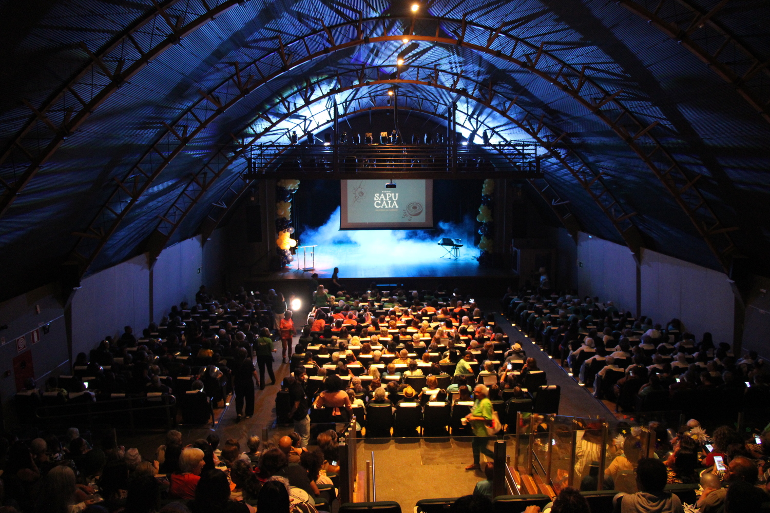 Encontro marca o reconhecimento e a valorização da arte e cultura das pessoas 60+ Fotografia panorâmica do interior de um teatro, registrada do fundo da plateia em direção ao palco. O espaço está lotado, com centenas de pessoas sentadas em poltronas organizadas em fileiras e algumas circulando pelos corredores. O público é formado, em grande parte, por pessoas idosas. O teatro possui teto alto e arqueado, com estrutura metálica aparente, iluminado em tons de azul. Ao fundo, o palco está iluminado por luzes azuis e apresenta um efeito de fumaça. Acima dele, uma tela exibe a inscrição “Sa