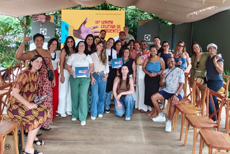 Menuuh! destaca experiências turísticas de Belo Horizonte na Semana Criativa de Tiradentes Grupo de pessoas lado a lado, algumas em pé, outras sentadas ou ajoelhadas, em frente ao painel da 10ª edição da Semana Criativa de Tiradentes
