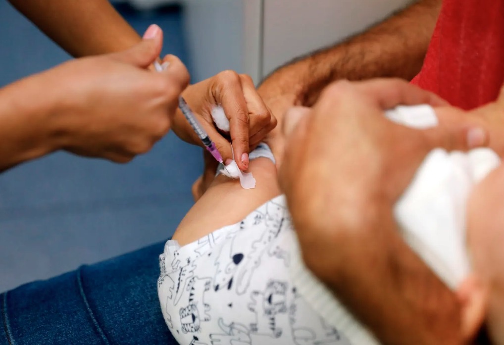 Imunização continua em BH após encerramento da Campanha de Multivacinação, na próxima segunda-feira (31) A foto mostra uma ação de vacinação em criança. A foto está em plano fechado e mostra as mãos de uma profissional de saúde aplicando uma vacina na coxa de um bebê, que é segurado no colo por um adulto.