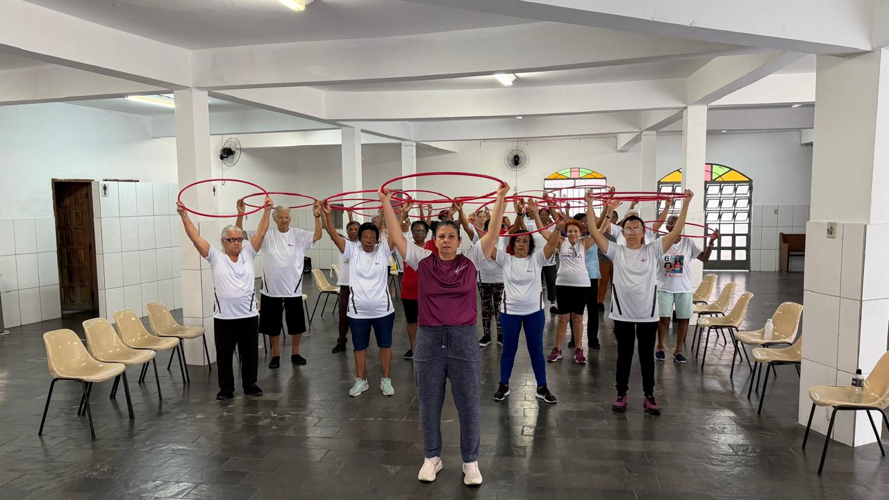 Em um salão amplo, a professora Jaqueline Munhoz à frente do grupo, de camiseta vinho, calça cinza e tênis branco, lidera a aula com os braços erguidos segurando um bambolê vermelho. Atrás dela, cerca de 20 alunas idosas de camiseta branca repetem o mesmo movimento, todas com bambolês vermelhos levantados acima da cabeça.