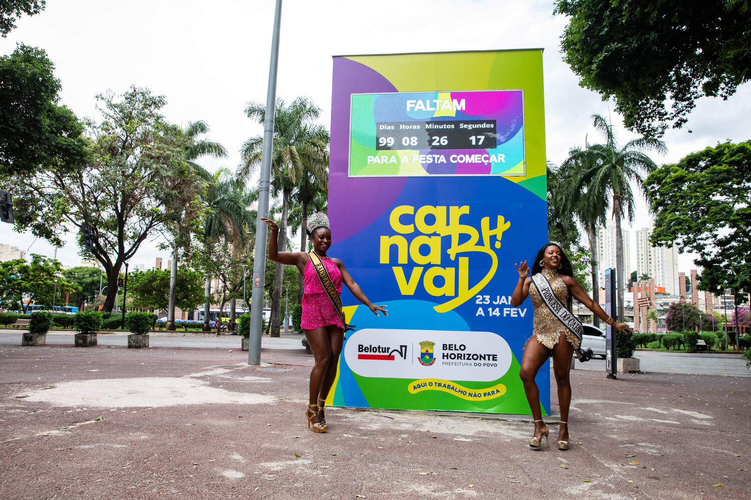 Totem marca contagem regressiva de 100 dias para o Carnaval de BH 2027 Totem colorido do Carnaval de Belo Horizonte, nas cores amarelo, verde, roxo e azul, com a marca “Carnaval de BH” e contador eletrônico que marca a contagem regressiva de 100 dias para a folia. Em frente ao totem duas mulheres, caracterizadas com roupa carnavalesca em posição de dança.
