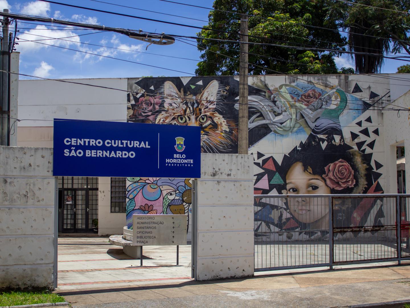 A foto mostra a fachada da entrada principal do Centro Cultural São Bernardo, capturada ao ar livre em um dia ensolarado, com céu azul parcialmente coberto por poucas nuvens. Primeiro plano: No centro, há um portão de entrada aberto com dois pilares brancos. Acima da passagem, há uma placa retangular azul com letras brancas indicando o nome "CENTRO CULTURAL SÃO BERNARDO" à esquerda, e à direita o brasão e a identificação "BELO HORIZONTE PREFEITURA". No chão do pátio interno, logo após a entrada, há uma pequena placa direcional de metal indicando setores como Auditório, Administração, Sanitários, Oficinas, Biblioteca e Praça. Segundo plano e paredes do prédio: A parede do edifício é decorada com coloridos grafites artísticos. No painel central superior, destaca-se a pintura detalhada do rosto de um gato com grandes olhos amarelados e orelhas pontudas, cercado por flores. À direita, um painel ilustra o rosto de uma criança negra com uma rosa cor-de-rosa no cabelo, cercada por formas geométricas e grafites abstratos. À esquerda, abaixo, há outras ilustrações coloridas com temas florais e da fauna. Detalhes adicionais: Um grande poste de concreto fica centralizado ao fundo, entre as artes, e a folhagem verde de árvores sobressai no topo superior direito da foto.