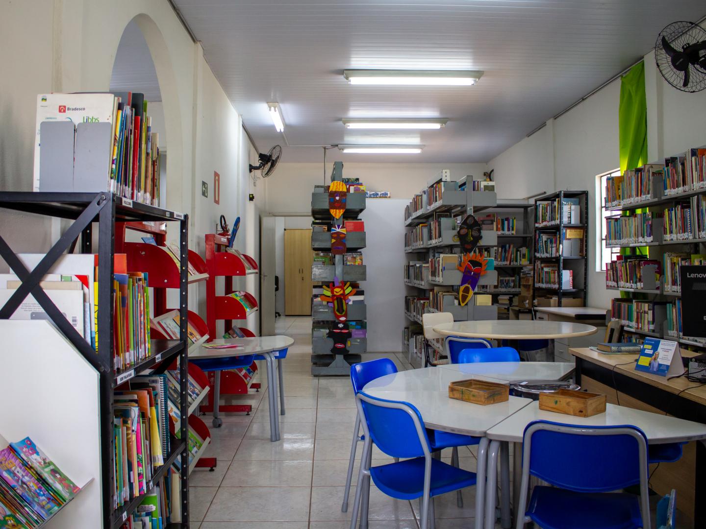 Fotografia em perspectiva de uma biblioteca bem iluminada. Em primeiro plano, do lado direito, há mesas brancas modulares dispostas juntas com várias cadeiras azuis de plástico ao seu redor. Sobre as mesas há pequenas caixas de madeira. Em toda a extensão da sala, do lado esquerdo, centro e fundo, há diversas estantes repletas de livros organizados. No corredor central e em uma das estantes, é possível ver máscaras decorativas coloridas de inspiração africana penduradas. O chão é de piso claro brilhante e o teto possui luminárias tubulares brancas acesas. As paredes são claras e o ambiente transmite uma sensação de organização e tranquilidade.
