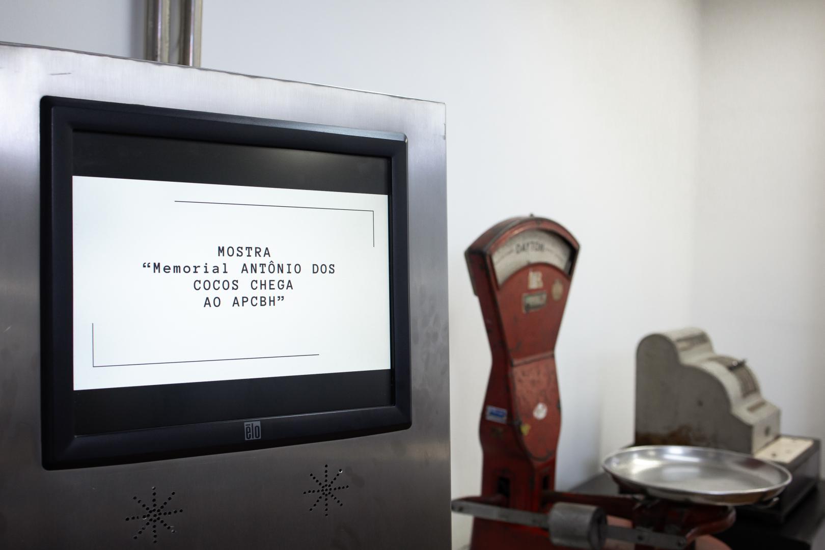 Uma imagem horizontal, em tons claros, mostra três equipamentos de medição do passado e do presente. À esquerda, em primeiro plano, uma estrutura de aço inoxidável emoldura uma tela de monitor com o logotipo "ēlo" na base central. A tela branca em preto exibe o seguinte texto: "MOSTRA 'Memorial ANTÔNIO DOS COCOS CHEGA AO APCBH'". As letras são maiúsculas e serifadas. À direita, ao fundo, fora de foco, estão uma balança mecânica de chão, vertical e de metal vermelho, e uma caixa registradora horizontal e de metal cinza. Uma bandeja de pesagem de metal circular está na base da balança mecânica. O fundo da imagem é uma parede branca e lisa.