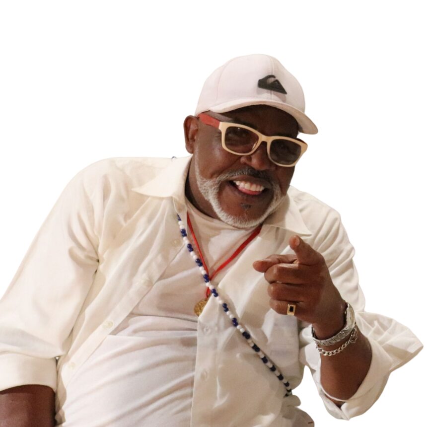 Fotografia em enquadramento de meio-corpo de um homem negro sorridente, recortado contra um fundo inteiramente branco. Ele está levemente inclinado para a frente, olhando para a câmera com uma expressão alegre e descontraída, sorrindo e mostrando a língua de forma brincalhona. Com a mão esquerda apontada em direção à câmera, ele veste uma camisa de botão branca aberta sobre uma camiseta também branca, boné claro com um pequeno logotipo escuro na frente, e óculos de armação bege clara com hastes vermelhas e lentes escuras. Ele possui barba e cavanhaque curtos e grisalhos. Usa acessórios como um colar de contas azuis, brancas e vermelhas, anel no dedo mínimo e pulseiras prateadas no pulso esquerdo.
