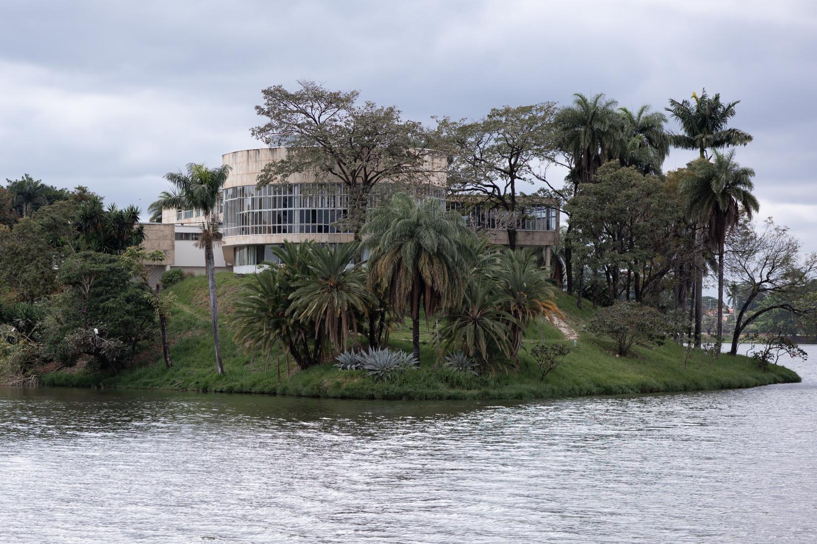 Fotografia em plano geral do Museu de Arte da Pampulha, situado na Lagoa da Pampulha, em Belo Horizonte. A edificação moderna e de formato cilíndrico, com paredes claras e grandes janelas de vidro em toda a sua extensão, fica posicionada no topo de uma elevação de terra coberta por grama verde. Ao redor do prédio e ao longo da colina, há uma vegetação densa composta por palmeiras, arbustos e árvores com copas frondosas. Em primeiro plano, a água da lagoa ocupa a parte inferior da imagem, refletindo de forma suave a luz do dia. O céu ao fundo está nublado e coberto por nuvens acinzentadas.