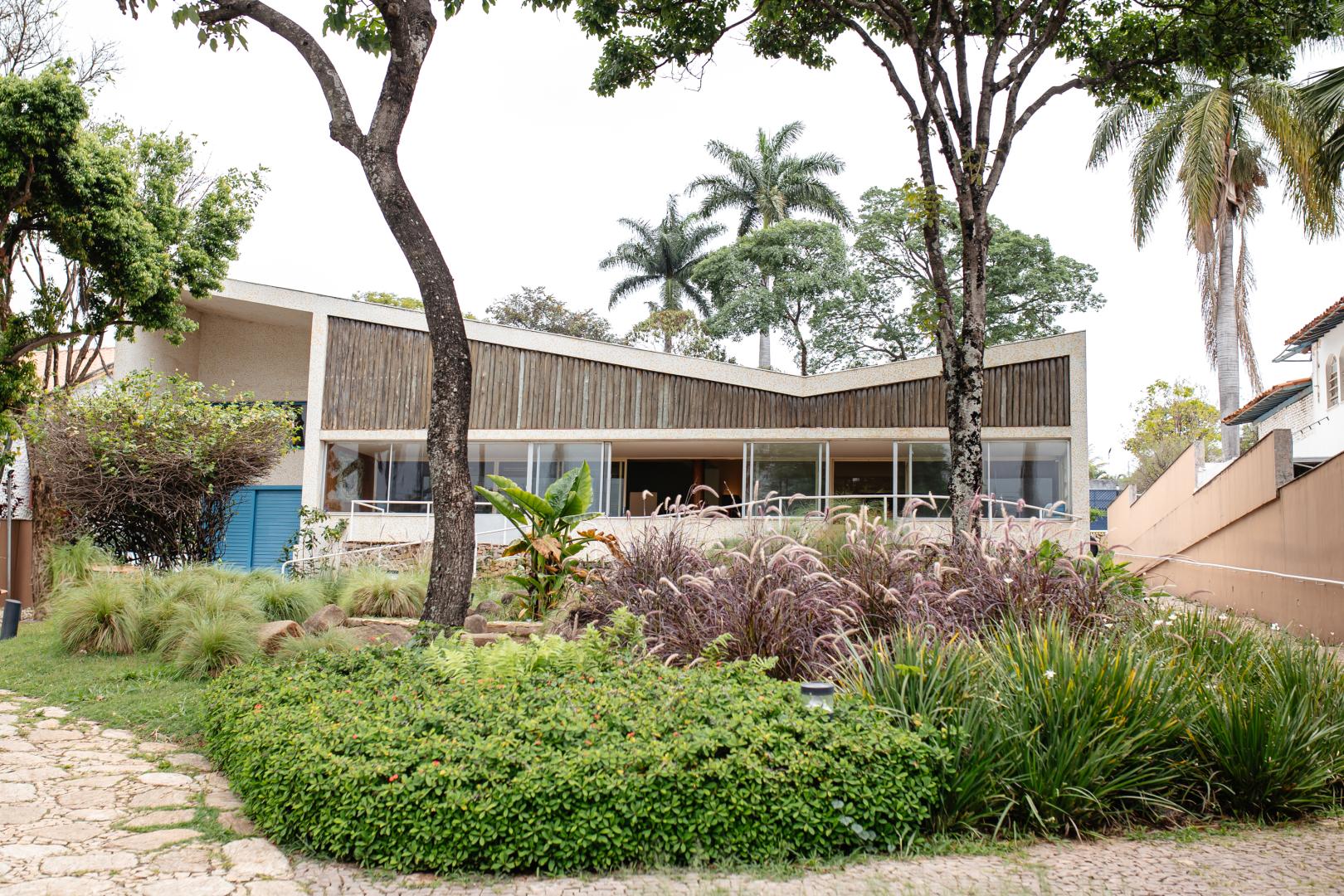 Fotografia ao ar livre, em plano médio, de uma casa modernista de dois andares com telhado em formato de borboleta, em um jardim exuberante. A fachada da casa é feita de vidro, madeira e concreto. O telhado é inclinado e as paredes são de vidro com painéis de madeira. O jardim é verde com árvores, arbustos e flores. Há uma calçada de pedra em primeiro plano. O céu está nublado. A imagem é bem iluminada e as cores são vibrantes. A composição é equilibrada e a foto é nítida.