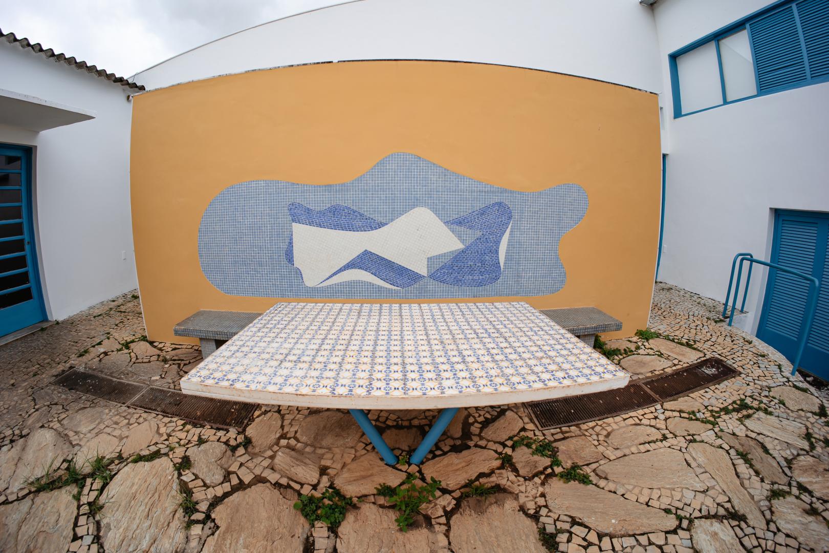 Fotografia em ângulo aberto de um pátio externo do Museu Casa Kubitschek, na Pampulha. Em primeiro plano, destaca-se uma grande mesa retangular revestida por pequenos azulejos decorados em tons de azul e branco, apoiada sobre um suporte metálico em V na cor azul. Atrás da mesa, há uma parede amarela de topo levemente curvo, ornada com um painel de pastilhas azuis e brancas com formas abstratas e onduladas. Ao lado e atrás da parede, observam-se estruturas brancas do edifício com portas e janelas com venezianas azuis. O chão do pátio é pavimentado com pedras irregulares de tom claro e calçada portuguesa.