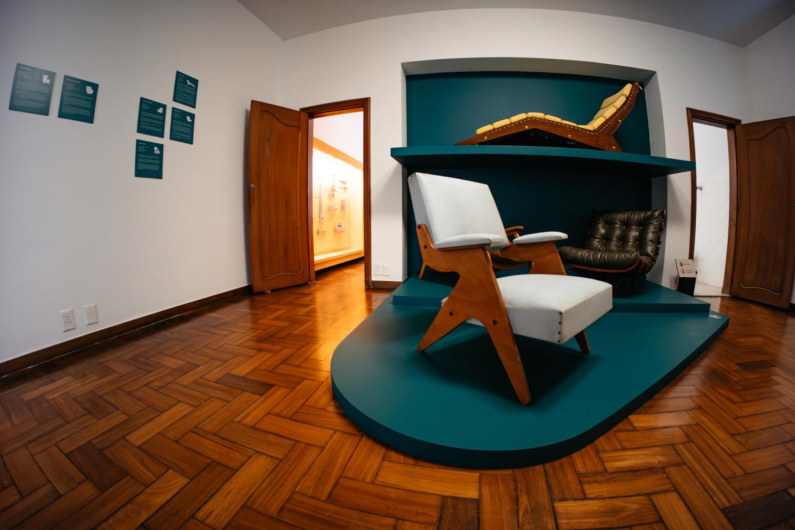 Fotografia em ângulo aberto do interior de uma sala de exposição com piso de taco de madeira em tom quente. Em primeiro plano, à direita, destaca-se uma plataforma baixa e curva na cor verde-petróleo, onde estão expostas peças de mobiliário de design. Sobre a plataforma, há uma poltrona de estofado claro com braços e pernas de madeira em formato geométrico. Ao fundo do expositor, dentro de um nicho da mesma cor verde-petróleo, vê-se uma poltrona baixa de couro escuro no nível do chão e, em uma prateleira suspensa acima dela, uma espreguiçadeira de madeira com estofado amarelo. Na parede branca à esquerda, há pequenos quadros explicativos na cor verde-petróleo alinhados na vertical e horizontal. Ao fundo, uma porta de madeira aberta revela outro ambiente iluminado e com itens expostos na parede.
