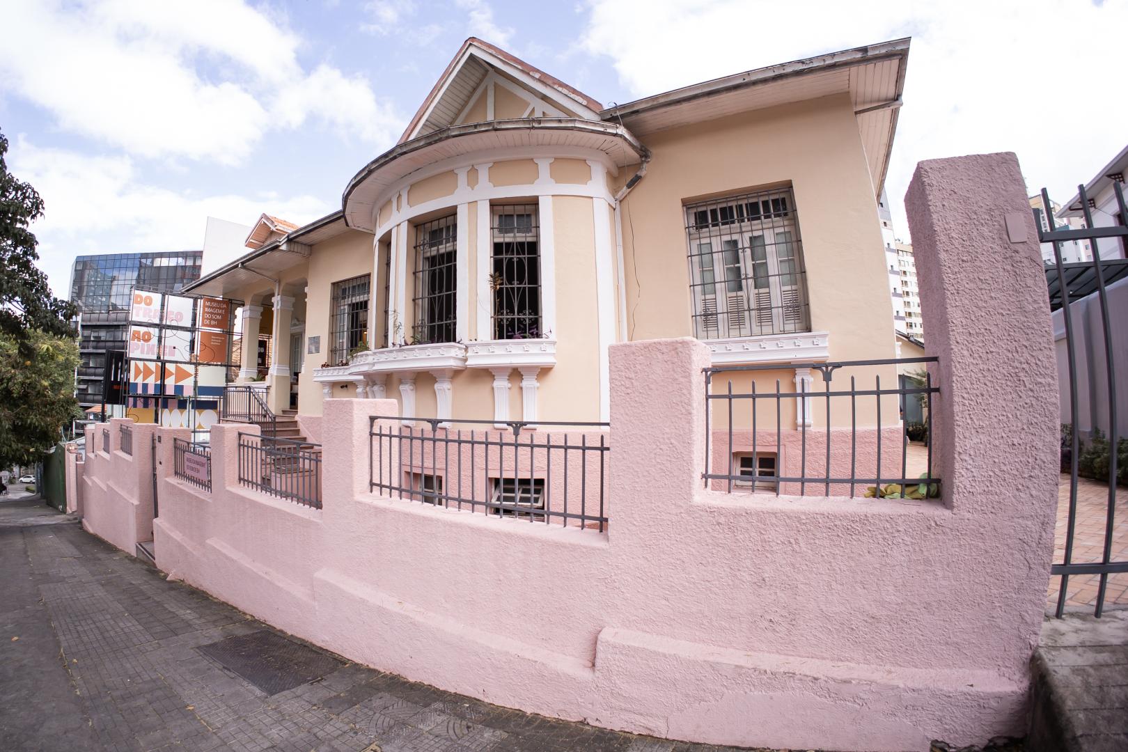 A fotografia mostra a fachada de um casarão antigo de estilo neoclássico e colonial, situado em uma rua inclinada. Casarão: A casa tem paredes em tom bege-claro com detalhes e frisos arquitetônicos em branco. Destaque para uma varanda arredondada central no segundo nível, com janelas em arco equipadas com grades de ferro e floreiras na base. Ao lado direito, há uma janela retangular maior com venezianas de madeira e grades. Muro e Portão: Na frente da casa, há um muro baixo de textura áspera pintado em tom rosa-antigo, intercalado por grades de ferro escuro. O muro acompanha a inclinação da calçada de paralelepípedos e concreto cinza à esquerda. Entrada e Entorno: À esquerda, escadas levam à varanda de entrada do imóvel. Ao fundo, no canto esquerdo, é possível ver um cartaz colorido de exposição e edifícios modernos sob um céu ensolarado com poucas nuvens brancas.
