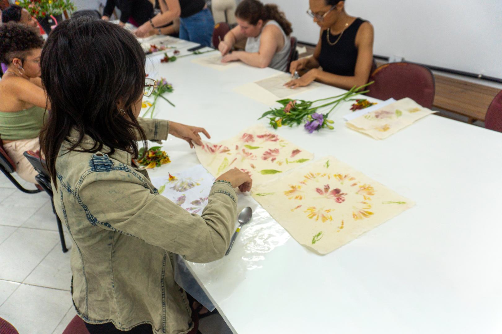 Uma fotografia em ambiente interno mostra um grupo de pessoas sentadas ao redor de uma grande mesa branca, participando de uma oficina de estamparia botânica. Primeiro plano: No canto esquerdo, em foco principal, uma mulher de cabelos escuros e jaqueta jeans clara está sentada de costas inclinadas para a mesa. Ela manuseia pedaços de tecido claro com estampas feitas a partir de folhas e pétalas de flores nas cores rosa, amarelo e verde. Sobre a mesa, ao lado do tecido, há uma colher de metal. Segundo plano: Ao longo da mesa branca, outras pessoas estão concentradas em suas atividades. Há ramos de flores coloridas (como astromélias roxas e rosa) espalhados sobre a superfície, além de papéis e materiais de trabalho. Ambiente: O espaço é bem iluminado, com paredes brancas ao fundo e piso claro de cerâmica.