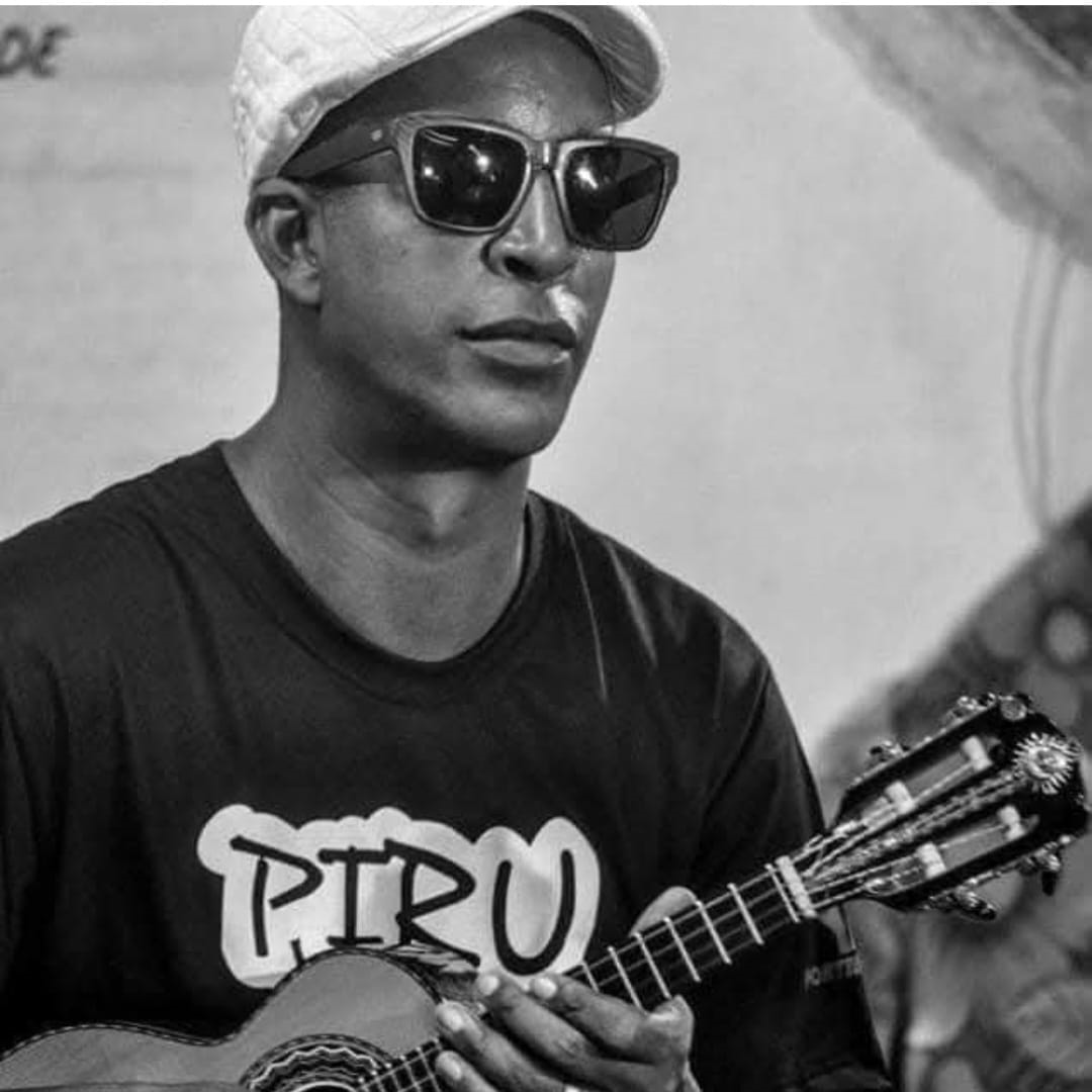 Fotografia em preto e branco de um homem negro do peito para cima, posicionado levemente de perfil voltado para a direita. Ele usa óculos escuros de armação quadrada, um boné ou boina clara de tom claro e uma camiseta escura com a palavra "PIRU" estampada em letras estilizadas em tom claro no peito. Ele segura e toca um cavaquinho ou instrumento de cordas pequeno, do qual é possível ver parte do corpo e o braço com a mão posicionada nas cordas. O fundo é claro e levemente desfocado.