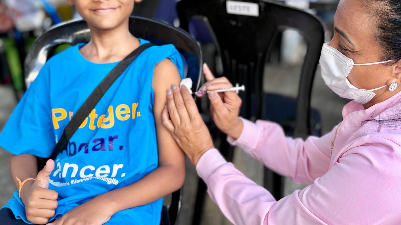 Prefeitura de BH aplicou cerca de 50 mil doses de imunizantes na Campanha de Multivacinação Criança sorridente, de camiseta azul, recebe uma injeção no braço enquanto faz sinal de positivo com a mão. Uma profissional de saúde, de máscara e blusa rosa, aplica a vacina. Cena de vacinação ao ar livre.