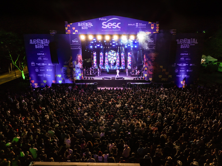 Multidão em frente ao palco. Estrutura grande e bem iluminada nas cores rosa e roxa, com palco no centro. 