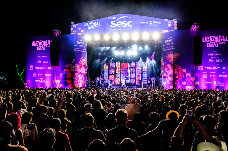 Multidão em frente ao palco. Estrutura grande e bem iluminada nas cores rosa e roxa, com palco no centro. 