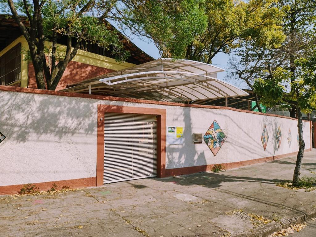 Foto da Foto da fachada da Escola Municipal Arthur Guimarães. A foto contém um muro branco com laranja e algumas pinturas, uma entrada para os(as) estudantes, um toldo em cima da entrada. Logo acima, árvores e, ao fundo, uma parte da escola.